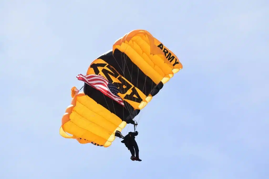 Paraquedista militar exibindo bandeira dos EUA durante salto com paraquedas amarelo e preto.