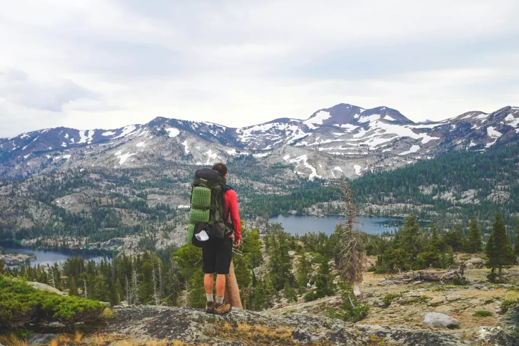 Homem com mochila de trekking apreciando paisagem de montanhas com neve e florestas ao fundo, em meio a uma trilha.