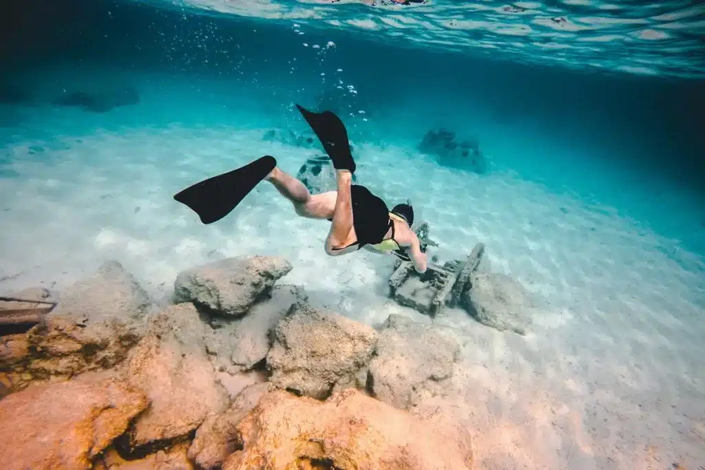 Mulher praticando mergulho livre com nadadeiras em águas cristalinas, explorando o fundo do mar com rochas e destroços.