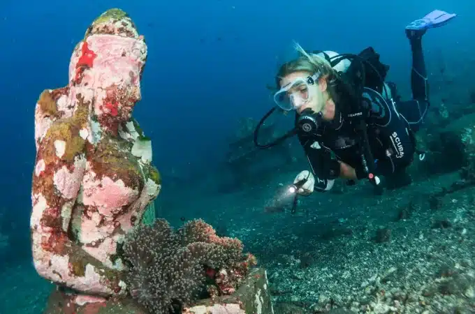 Mergulhadora com equipamento completo e lanterna explorando uma estátua submersa coberta por corais no fundo do mar.