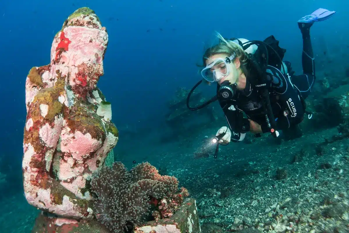 Mergulhadora com equipamento completo e lanterna explorando uma estátua submersa coberta por corais no fundo do mar.