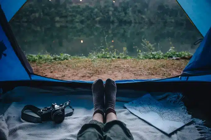 Vista de dentro de uma barraca de acampamento com pés de uma pessoa deitada, câmera analógica e mapa ao lado, com um lago e vegetação ao fundo.