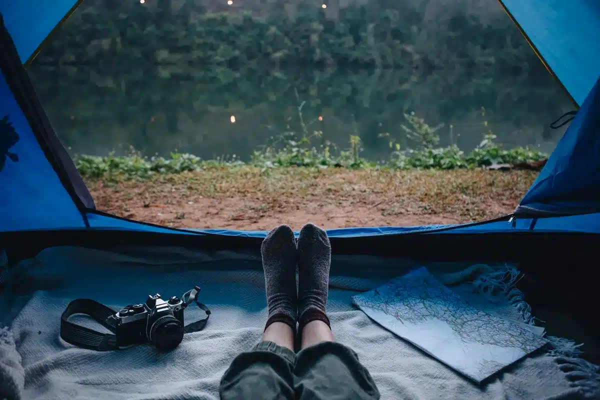 Vista de dentro de uma barraca de acampamento com pés de uma pessoa deitada, câmera analógica e mapa ao lado, com um lago e vegetação ao fundo.