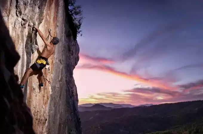 Homem praticando escalada em uma parede rochosa íngreme ao ar livre, com o céu ao fundo em tons de roxo, rosa e laranja durante o pôr do sol.