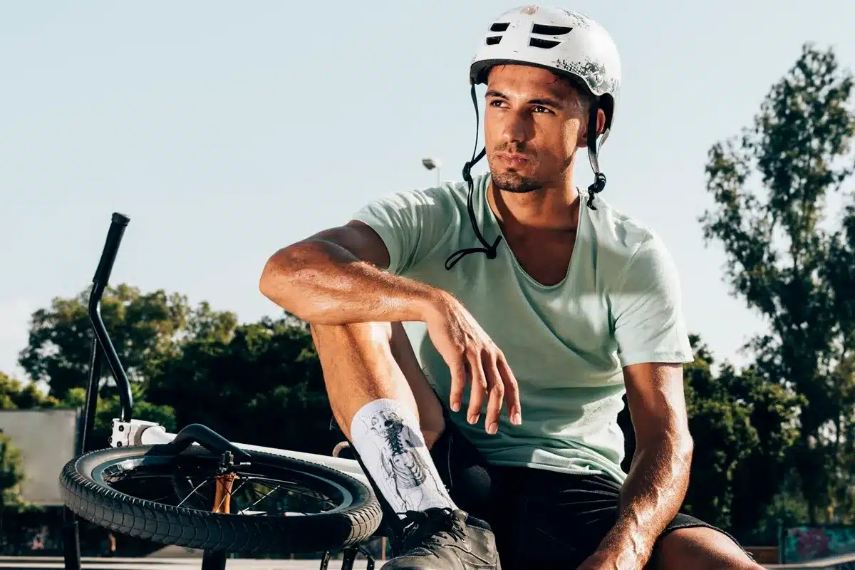 Homem com capacete de proteção branco, sentado ao lado de uma bicicleta BMX em um ambiente ao ar livre, com expressão pensativa. Ele veste uma camiseta verde clara e shorts escuros.