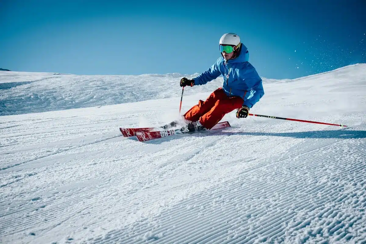 Esquiador descendo uma pista de neve com roupa azul e calça vermelha em um dia ensolarado com céu azul.