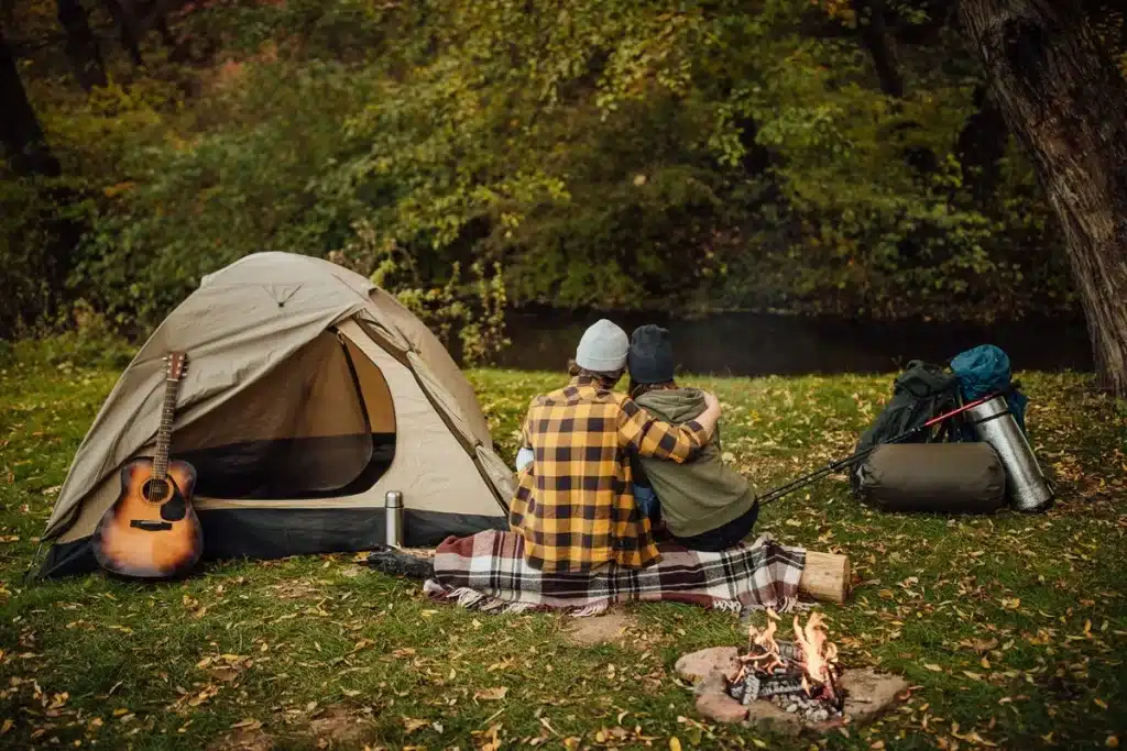 Casal abraçado sentado em frente a uma barraca de camping ao lado de uma fogueira acesa, com violão e equipamentos de camping em meio à natureza.