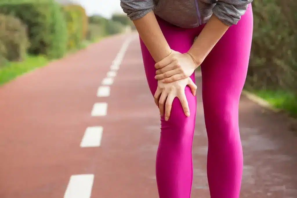 Pessoa segurando o joelho em uma pista ao ar livre, vestindo calça esportiva rosa e moletom cinza, indicando desconforto ou lesão.