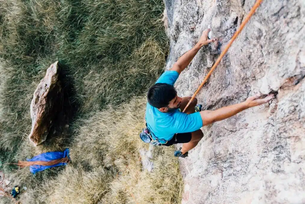 Homem vestindo camiseta azul e shorts escuros, subindo uma parede rochosa com equipamento de segurança e corda laranja. Abaixo, vegetação densa e uma mochila azul com cordas organizadas.