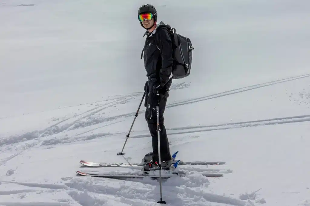 Esquiador em pé na neve com traje preto, capacete e óculos de proteção espelhados, carregando uma mochila e segurando bastões de esqui.