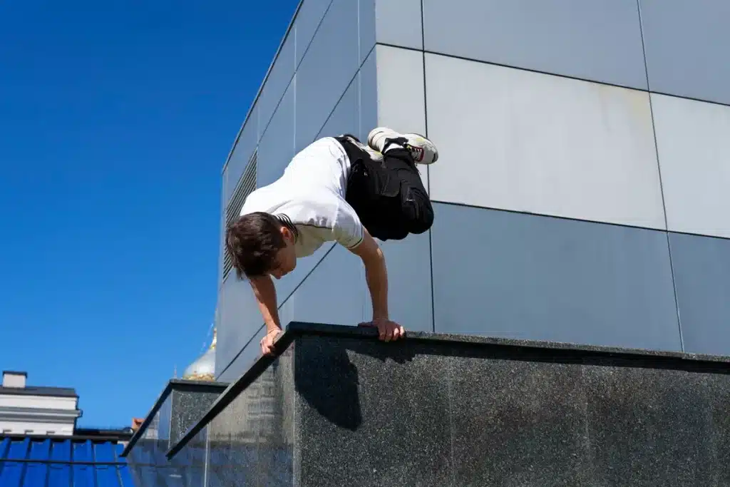 Homem praticando parkour em um prédio, realizando uma manobra de equilíbrio e força contra um céu azul claro.