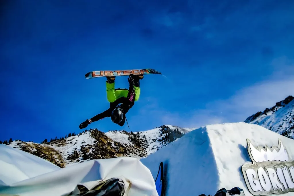 Atleta realizando uma manobra aérea de snowboard com prancha colorida em pista de neve com céu azul ao fundo.