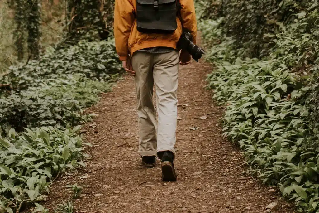 Pessoa com jaqueta amarela e mochila preta caminhando em uma trilha rodeada por vegetação verde, segurando uma câmera profissional.