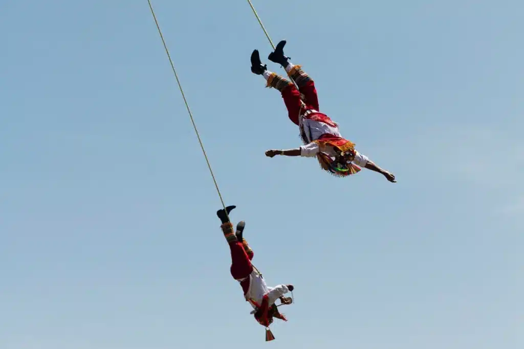 Dois dançarinos vestidos com trajes tradicionais mexicanos realizando a dança dos Voladores de Papantla, pendurados em cordas contra o céu azul.