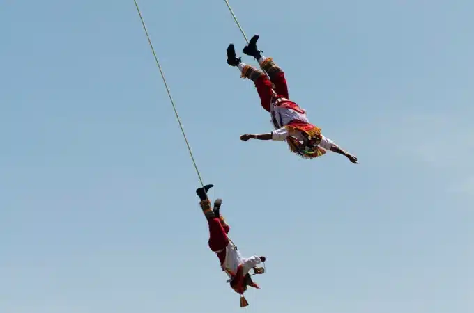 Dois dançarinos vestidos com trajes tradicionais mexicanos realizando a dança dos Voladores de Papantla, pendurados em cordas contra o céu azul.