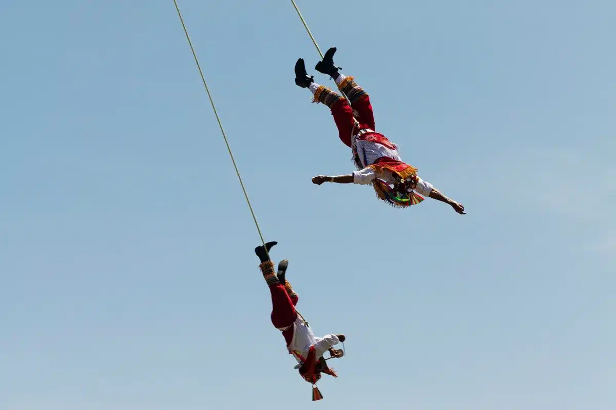 Dois dançarinos vestidos com trajes tradicionais mexicanos realizando a dança dos Voladores de Papantla, pendurados em cordas contra o céu azul.