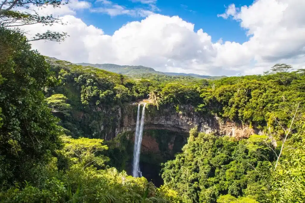 Cachoeira em um parque nacional cercada por uma densa floresta verde com céu azul e nuvens ao fundo.