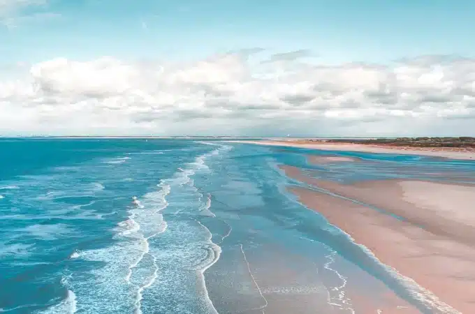 Vista panorâmica de uma praia deserta com areia clara, mar azul e pequenas ondas quebrando suavemente sob um céu com nuvens brancas.