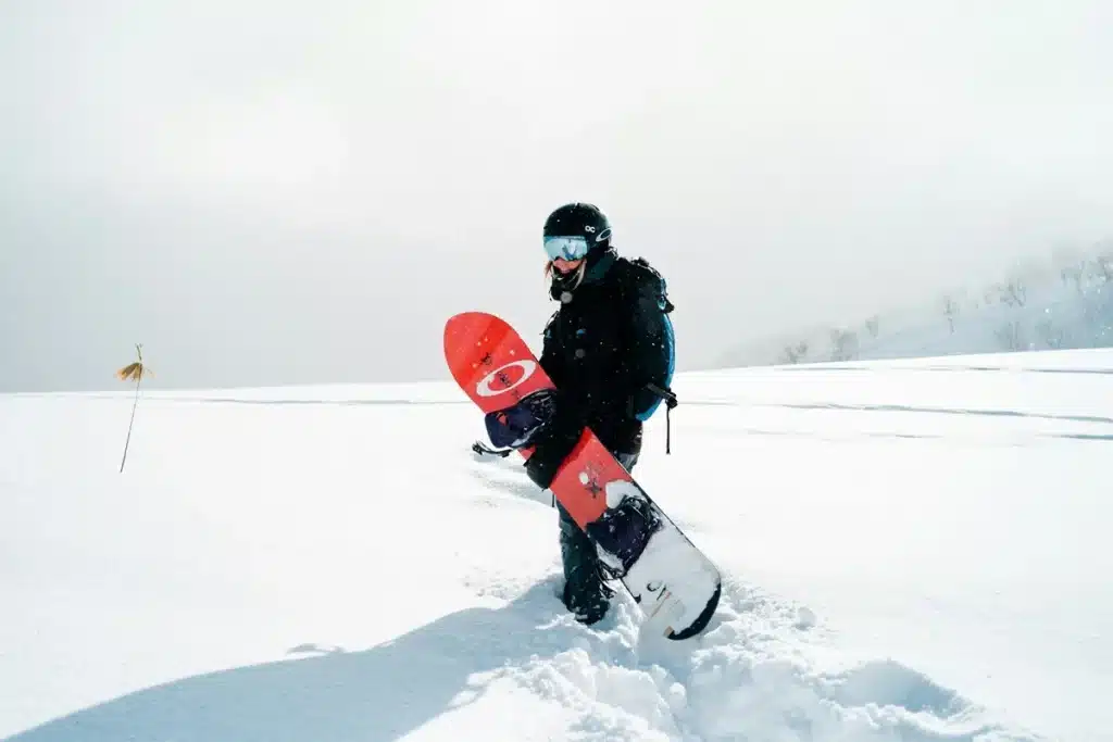 Pessoa praticando snowboard em uma paisagem coberta de neve, segurando a prancha vermelha com design moderno, equipada com capacete e óculos de proteção.