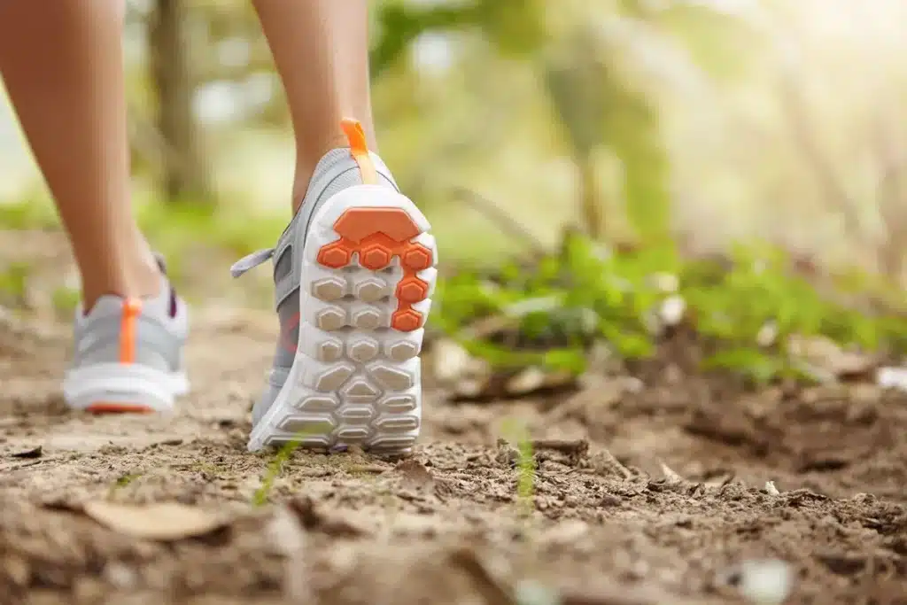 Close de pés com tênis de trilha cinza e detalhes laranja em uma trilha de terra com vegetação ao fundo.