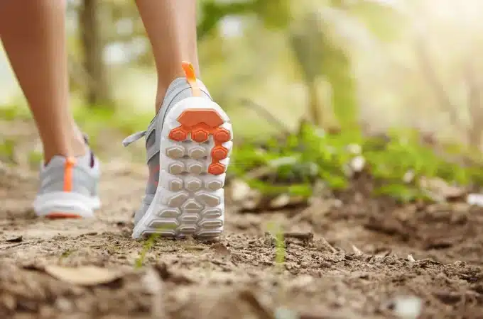 Close de pés com tênis de trilha cinza e detalhes laranja em uma trilha de terra com vegetação ao fundo.