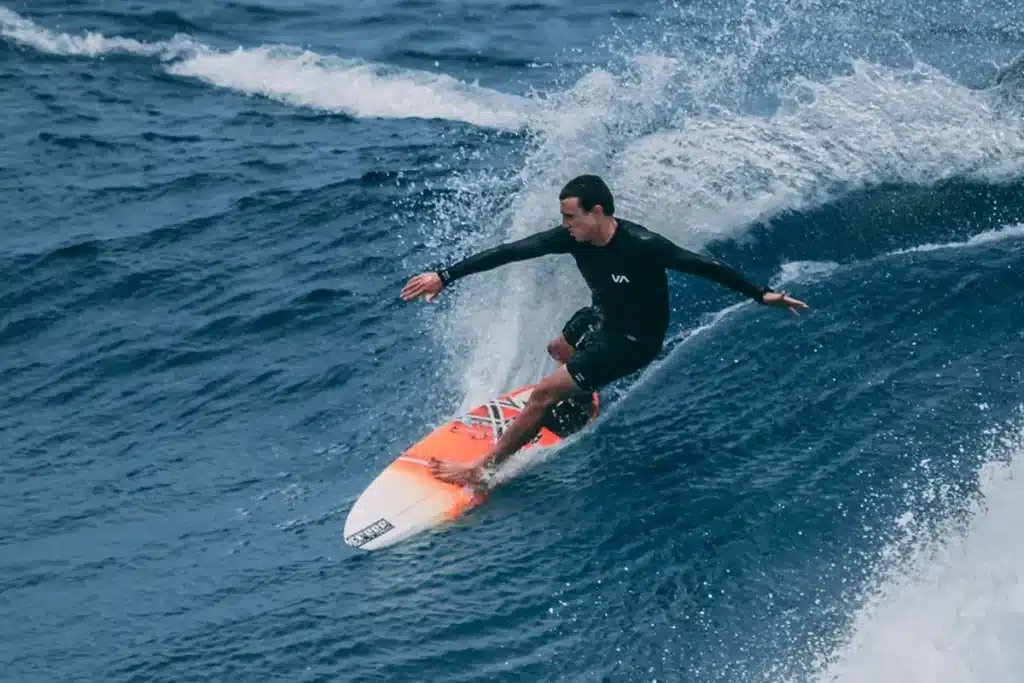 Surfista vestindo roupa preta deslizando sobre uma onda azul intensa com sua prancha laranja e branca.