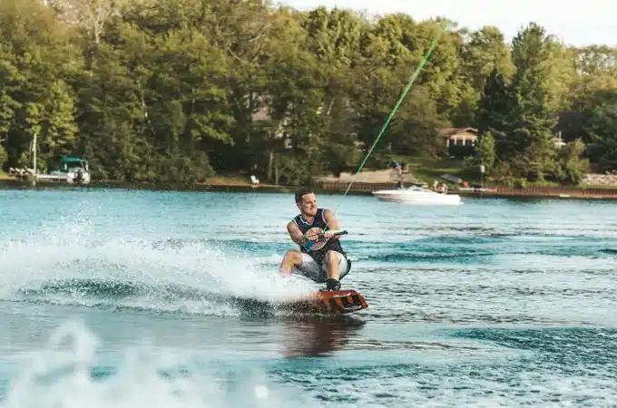 Homem praticando wakeboard em um lago, puxado por uma corda, gerando ondas e respingos de água ao deslizar sobre a superfície.