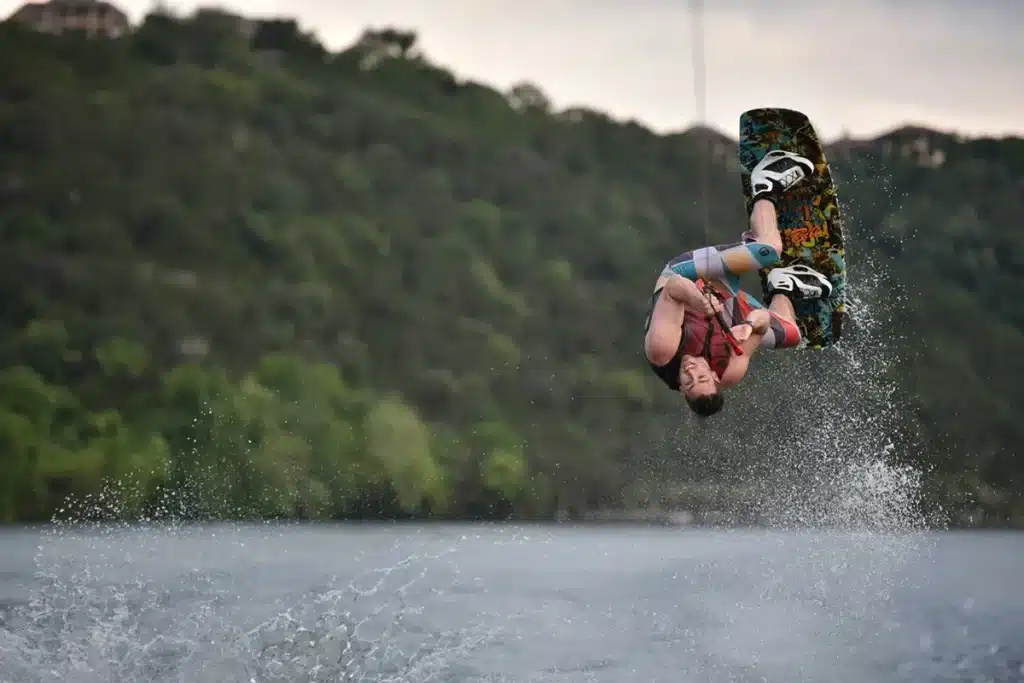 Atleta de wakeboard realizando uma manobra aérea impressionante, segurando a corda enquanto gira de cabeça para baixo acima da água.