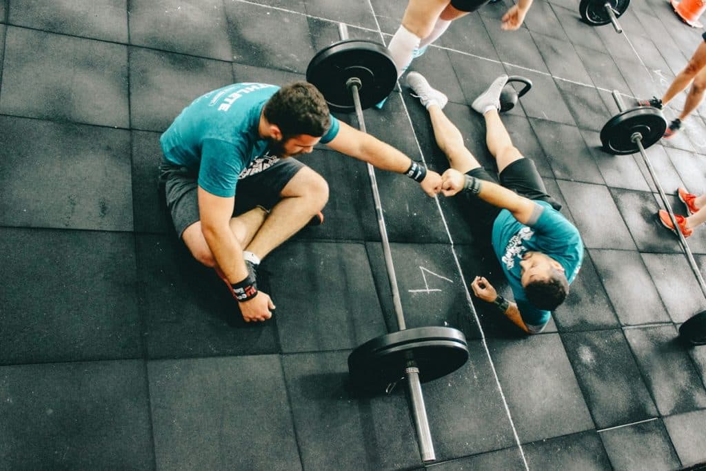 Dois homens em um treino de cross training, sentados no chão e cumprimentando-se após um exercício intenso com barra.