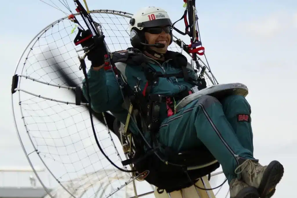 Piloto de paramotor sorrindo durante o voo, equipado com capacete, óculos de proteção e traje esportivo.