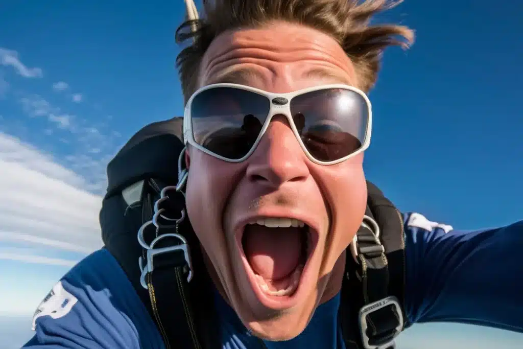 Homem sorrindo e gritando de emoção durante um salto de paraquedas, com óculos escuros e traje esportivo em pleno voo.