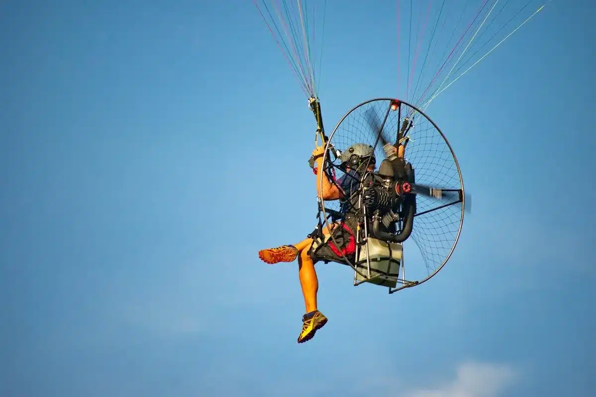 Piloto de paramotor voando no céu azul com equipamento motorizado e parapente colorido.