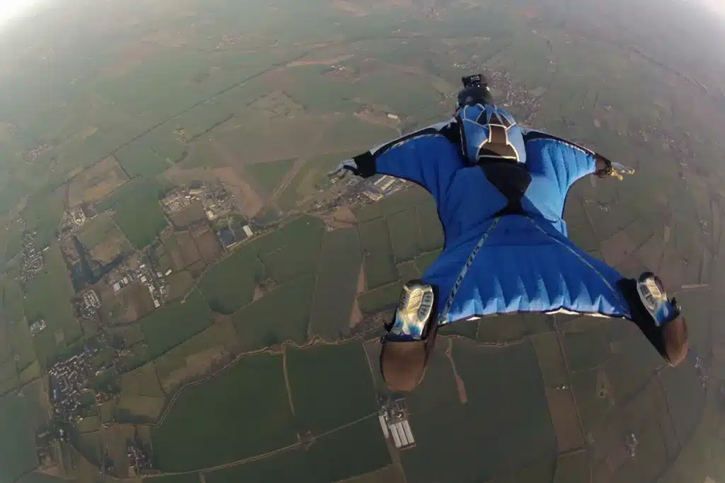 Praticante de wingsuit voando a grande altitude sobre uma paisagem rural, usando traje azul aerodinâmico para planar.